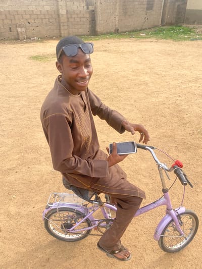A young man is depicted sitting on a light purple bicycle on a sandy, open ground, smiling towards the camera. He is wearing a brown long-sleeved tunic-style shirt with intricate gold embroidery and matching trousers, along with dark sandals. A pair of dark sunglasses rests on top of his head. He holds a white-cased smartphone in his right hand, looking at the screen, while his left hand rests near the bicycle's handlebars.

The bicycle is small, possibly a child's or compact adult's bike, and features a chrome rear rack. The ground is primarily sandy, with a strip of green grass and some litter visible along the left side of the frame. In the background, a light-colored block wall, typical of construction in some regions, stretches horizontally, indicating an outdoor, possibly undeveloped or residential area. The scene is brightly lit by daylight, suggesting a clear day. The man's smile conveys a relaxed and happy mood. Text is faintly visible on the bicycle frame, with "SPORT" and possibly "JAY" or "BIKE" discernible, though not fully legible. The smartphone screen shows dark icons or a dark interface. The setting is consistent with a location in Tilde, Nigeria.
