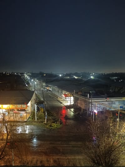 An elevated forensic view depicts a nighttime urban scene in Khasanbay, Uzbekistan, following a rain event, evidenced by numerous reflections on wet surfaces. The dark, clear sky occupies the upper portion of the frame. A primary asphalt road runs from the lower-left foreground towards the center-right midground, where it curves right and extends into the distance, illuminated by multiple streetlights and reflecting light sources.

On the immediate left, a single-story building with a dark roof displays a brightly illuminated, yellow-orange interior, possibly a commercial establishment. Next to it, a white awning-like structure is partially visible. Further down the street on the left, additional dark-roofed buildings with some illuminated windows are present.

On the right side of the main road, a long, low-rise building features a partially obscured red and white sign, possibly reading "BITERA." Adjacent to this, another building bears a large blue sign with Cyrillic text, including "ГИДРОКОМПЛЕКТ", "ПЕСТИЦИДЫ", and "ГИДРОСТРОЙ," indicating a building supply or similar retail outlet. Red and pink neon signage is also visible on this side.

Multiple utility poles and overhead power lines crisscross the mid-to-lower frame. Bare-branched deciduous trees, indicative of late autumn or winter, are prominent in the lower left and lower right foreground, partially obscuring the view. A red reflection is visible on the wet pavement near the "BITERA" sign. In the distance along the main road, at least two indistinct white vehicles are present. No individuals are visible. The environment is characterized by artificial light reflecting off wet surfaces, creating a shimmering effect.
