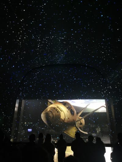 A low-angle shot captures a large screen displaying a space shuttle in a planetarium setting. Above, a simulated night sky is filled with countless stars. Silhouetted figures of people stand in the foreground, their attention focused on the screen. The scene is dimly lit, highlighting the bright display and the starry "sky" above.