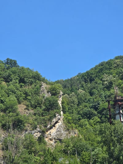 The image shows a lush green mountainside covered in trees, with a rocky cliff face. The sky above is a clear, bright blue. On the right side of the image, there are remnants of an industrial structure, appearing to be a part of the former Alumina factory in Zvornik, Bosnia and Herzegovina. The structure consists of tall metal pipes and platforms, weathered and partially covered by vegetation. The foreground is dominated by dense foliage and some bare tree branches. The overall scene depicts a natural landscape intertwined with industrial ruins.