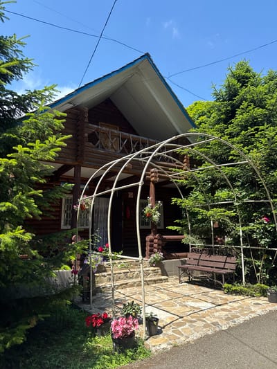 The image depicts a rustic wooden house with a blue-trimmed roof, set amidst lush green trees. In front of the house, a white arched trellis stands over a stone pathway. A brown metal bench with ornate scrollwork is placed to the right of the trellis, partially shaded by the trees. Potted flowers in shades of red and pink are visible near the entrance and along the pathway. The sky above is clear and blue. This scene is located in the city of Goryachy Klyuch, Russia.