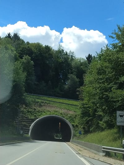 The image shows a road leading towards a tunnel entrance, situated in a wooded area under a bright blue sky with fluffy white clouds. A white lane divider is visible on the asphalt road. To the right of the road, a concrete barrier and a guardrail are present, followed by a grassy embankment and lush green trees. A traffic sign with the German word "Licht" (light) and an icon of headlights is posted on the right side. To the left of the tunnel entrance, a steep, terraced embankment covered in vegetation is visible. The tunnel is a dark, arched structure. The scene appears to be located in or near Hutthurm, Germany, given the presence of the German traffic sign.