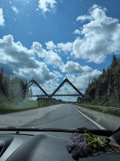 The image depicts a first-person perspective from inside a vehicle, looking forward through the windshield onto a highway under a partly cloudy blue sky. The prominent structure in the midground is the Kirjola Bridge in Heinola, Finland, identifiable by its two inverted 'V'-shaped brown pylons from which suspension cables extend to support the roadway.

The asphalt highway features white lane markings and a metal guardrail along the right side. In the distance, other vehicles are visible on the road. Dense green trees and foliage line both sides of the highway, with a significant rocky cliff face covered in sparse vegetation present on the immediate right. The sky is bright blue, populated by large, fluffy white cumulus clouds, with some sun glare and lens flare visible in the upper sections. On the vehicle's dashboard, located on the right side in the foreground, rests a small bouquet of purple flowers with green leaves.