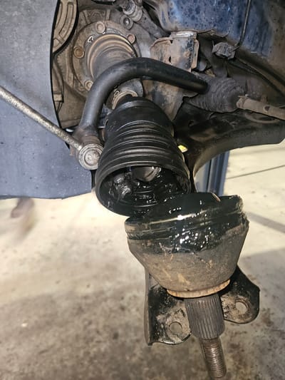 A close-up, ground-level view of a damaged automotive suspension component, specifically a torn CV (Constant Velocity) joint boot. The black rubber boot is ripped, exposing its internal grease and parts. The boot is connected to a black metal arm above and a metallic component with a ribbed shaft below. The background appears to be a concrete floor, suggesting an indoor setting such as a garage or workshop. No people or city-specific landmarks are visible. The lighting is neutral.
