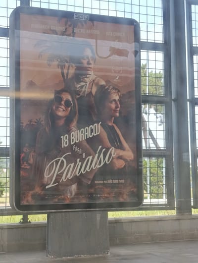 The image shows a movie poster displayed on a light box, likely at a transportation hub or public space in Palmela, Portugal, given the context. The poster features three women. The central figure is a woman looking forward with a scarf around her neck. To her left, a woman with sunglasses smiles and looks to the side. To her right, a woman with short hair gazes away from the camera. The poster's title is prominently displayed as "18 BURRACOS PARA O Paraíso" in large, stylized text. Below the title, the text "REALIZADO POR JOÃO NUNO PINTO" is visible. The background of the poster depicts a desert landscape with palm trees and mountains, rendered in warm, sepia tones. The light box is framed by a dark metal border and is supported by a dark gray structure, suggesting it is part of an outdoor or semi-outdoor installation. The overall scene is illuminated by natural light, with a grid-like structure of windows and the sky visible through the background, indicating an indoor setting with large windows looking out onto an outdoor area.