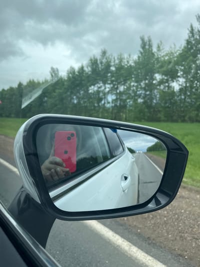 An image captured from within a vehicle shows a view through the driver's side window and into the side mirror. The prominent white side mirror, partially adorned with water droplets, reflects a person's hands holding a bright red Apple iPhone with dual camera lenses. The person's fingers exhibit light-colored nail polish. The reflection also reveals a portion of the white car body and a segment of the asphalt road receding into the distance, with what appears to be another vehicle or light source far down the road. Beyond the car, a clear view through the window and in the mirror displays a rural landscape under an overcast sky. A paved road with a white line borders a vibrant green field, which is further bordered by a dense line of green trees. The scene is located in Bizyaki, Russia.