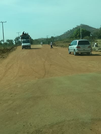 The image depicts an unpaved, reddish-brown dirt road in Jibam, Nigeria, under a clear, light-blue sky. Tire tracks are visible across the dusty road surface. A white delivery van, heavily loaded with cargo and multiple passengers seated atop its roof, travels away from the viewer on the left side of the road, generating a dust trail. Further down the road, a motorcycle with a single rider also moves away from the viewer, creating a smaller dust cloud. A pedestrian wearing a light-colored shirt is observed walking from right to left across the mid-ground of the road. On the right, a silver minivan is stationary, oriented away from the camera. The background features a gentle hill with sparse vegetation and multiple utility poles supporting overhead lines. Additional indistinct human figures are present further down the road.