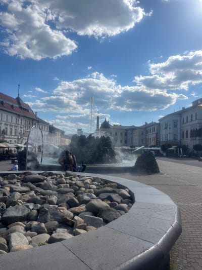 The image captures a vibrant outdoor scene in Banská Bystrica, Slovakia, on a bright, sunny day with scattered clouds. The perspective is from a fountain area, looking towards a street lined with elegant European-style buildings.
The foreground is dominated by a large, decorative fountain filled with rounded river stones. Water is actively spraying from the fountain, creating a misty effect. Two individuals are seated on the edge of the fountain, one appearing to be a woman in a patterned dress. A man in a white cap is partially visible near the bottom of the frame.
The background features a row of multi-story buildings with distinct architectural details, including arched windows and ornate facades, typical of a historic European town square. Some buildings show signage, though it is not legible. Outdoor seating and umbrellas suggest the presence of cafes or restaurants. The sky is a brilliant blue, with fluffy white clouds adding depth and texture.
The overall impression is of a lively public space enjoying pleasant weather. The light suggests it is daytime, likely late morning or afternoon, given the bright illumination. The scene evokes a sense of leisure and community in a picturesque setting.