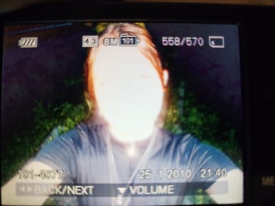 A still image displays the screen of a digital camera showing a photograph taken outdoors. The photograph features a person, appearing to be female, with long, light brown hair. She is wearing a light blue shirt. The background is a blur of green foliage, suggesting an outdoor setting with vegetation. Overlaid on the image are several icons and text indicators from the camera's interface. At the top left, a battery icon shows a low charge level. To its right, text reads "4.3 8M 101". Further to the right, "558/570" is displayed above another battery icon. At the bottom left, "101-4077" is visible above a "BACK/NEXT" control. At the bottom right, the date "25 1 2010" and time "21:40" are displayed, along with a "VOLUME" control. The location is identified as Pokrovka, Russia.