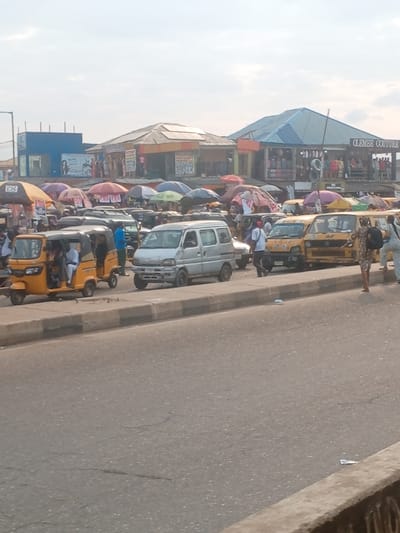 The image depicts a busy street and market scene in Abule Egba, Nigeria, under daylight conditions with a partially clouded sky.

In the foreground, a grey asphalt road with minor surface cracks extends across the lower portion of the frame. A grey concrete curb separates the road from an elevated pedestrian or parking area. A lighter grey concrete barrier is visible in the bottom right corner, partially obscuring the immediate foreground.

The midground is dominated by stationary or slow-moving vehicles and pedestrians. From left to right:
*   A yellow three-wheeled auto-rickshaw (Keke Napep) is positioned near the curb, with two occupants visible: one in white attire and another in blue.
*   Behind the auto-rickshaw, a silver compact van, with visible damage to its front bumper, is stopped.
*   Further right, a line of at least three yellow commercial minibuses are parked or stopped along the elevated curb.
*   Pedestrians are moving through the scene. A male subject wearing a white t-shirt and dark trousers walks from right to left between the silver van and the yellow minibuses. A female subject wearing a patterned dress and a dark backpack walks from right to left alongside the yellow minibuses. Other individuals are partially visible among the vehicles and stalls.
*   Behind the line of vehicles, a dense arrangement of market stalls is present, covered by numerous umbrellas of various colors including red, blue, purple, green, and black. Signs reading "POS" are discernible on some stalls.

The background features multiple commercial buildings.
*   On the far left, a blue building with a flat roof is visible.
*   Towards the center-left, a building with an orange corrugated metal roof features solar panels and a sign reading "PHOTOCopy."
*   To the right, a larger building with a blue corrugated metal roof displays a prominent sign reading "CLEMBE COUTURE" and shows clothing items on display.