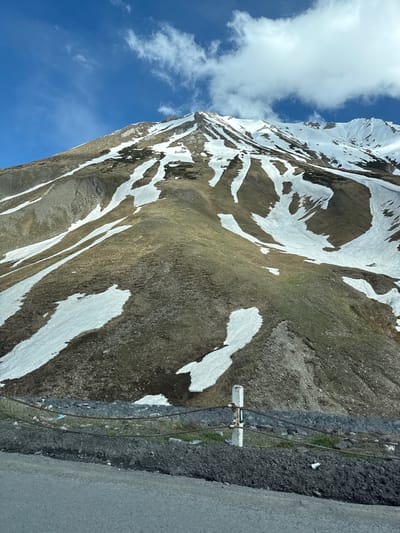 A sharply inclined mountain slope dominates the frame. The landscape is a mix of brown earth and lingering snow patches. A small, utility structure with a vertical and horizontal white beam and some wires is near the side of the mountain. To the right, there is a roadway with a gray concrete barrier. The sky is blue with a few clouds. This is near Almasiani, Georgia.