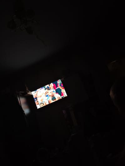 In a dimly lit room, likely a living room, a television screen is visible, displaying a broadcast. The screen shows a crowd of people, some wearing white and blue attire, appearing to be at an event or sporting match. A score overlay can be seen on the bottom left of the screen, and a logo in the top right corner of the broadcast. The room's ceiling features a chandelier with multiple lamps. The overall scene suggests a relaxed evening indoors, possibly watching a game with others, though no other individuals are clearly depicted. The location is specified as Грачёвка, Russia.