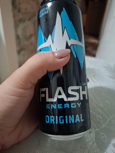 A hand with light pink nail polish is holding a black energy drink can. The can has a design with blue and white jagged lines and text that reads "FLASH ENERGY ORIGINAL". The background is blurred and appears to be an indoor setting with some textured wallpaper and a white surface. There are water droplets on the can, suggesting it is cold. There is no information about the location in Novlyanka, Russia.