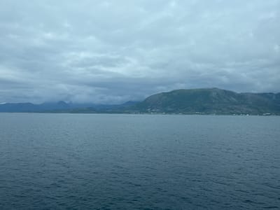 This is a wide-angle, eye-level shot of a body of water with mountains in the background. The water is a dark blue-gray and appears slightly choppy. The sky is overcast with heavy, gray clouds. In the distance, a range of green mountains slopes down to the water, with a small village or settlement visible along the shoreline. The scene is likely from the municipality of Lødingen in Norway, given the characteristic coastal mountain landscape. There are no people or discernible objects in the foreground or midground.