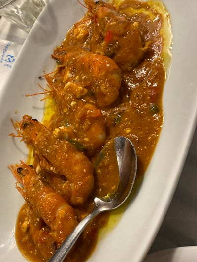 A white rectangular plate holds a serving of approximately seven whole cooked shrimp, heads and tails intact, coated in a thick, glossy, orange-brown sauce. The sauce contains visible specks of red and green vegetables, and numerous small, white, crumbled pieces, possibly cheese. A silver spoon with a slightly ornate handle is partially submerged in the sauce, resting on the plate. In the upper left background, a clear glass tumbler is partially visible. A white surface with a faint blue pattern, possibly a logo resembling "Ξ2" or similar characters on a napkin or tablecloth, is present in the background. The scene is located in a dining establishment in Megalochori, Greece.