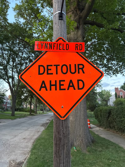An orange diamond-shaped road sign stating "DETOUR AHEAD" is mounted on a wooden pole. Above the detour sign, a smaller orange rectangular sign reads "LYNNFIELD RD." The sign is located on a residential street in Shaker Heights, United States of America. Trees line the street, and houses are visible in the background. The sky is overcast.