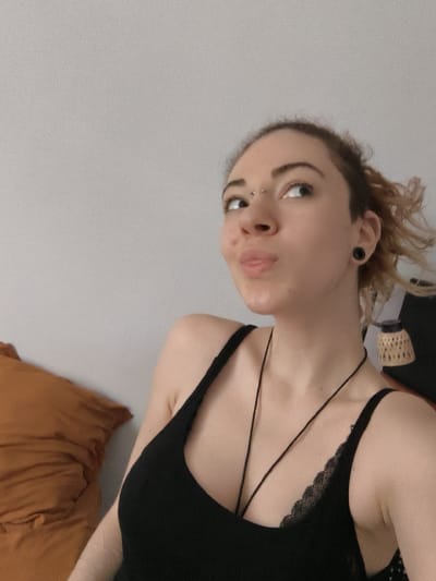 The image displays a female individual situated indoors in La Madeleine, France. The subject, positioned centrally and to the right, has fair skin, light-colored eyes, and light brown hair styled with some strands visible around her head. A bridge piercing with two silver studs is present on her nose. Her left earlobe (viewer's right) features a black circular ear gauge. Her lips are pursed and slightly puckered, with her tongue partially visible. Her gaze is directed upwards and towards the right of the frame. She is attired in a black tank top with thin straps. A black lace bra is visible beneath the tank top on her right side (viewer's left). A thin black cord necklace hangs around her neck, extending over her chest. The background consists predominantly of a light gray wall. To the lower left, a portion of a large orange or brown object, resembling a pillow or folded textile, is visible. To the upper right, behind the subject's right shoulder, a light-colored decorative object with a woven or patterned shade is partially visible.