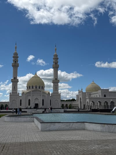 In the city of Privolzhsky, Russia, a large, white mosque with a golden dome stands under a bright blue sky with scattered white clouds. The mosque has two tall minarets flanking the central structure, which also features an arcade with arches. In front of the mosque, there is a paved courtyard and a shallow, blue-tiled reflecting pool with tiered marble edges. Several people are visible in the courtyard, some walking and others standing near the mosque entrance and by the pool. The area appears to be a public space, possibly a tourist attraction or a place of worship. The overall scene is serene and picturesque.