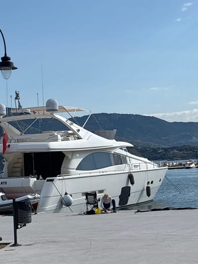 In Νέα Ηρακλείτσα, Greece, a large white yacht is moored alongside a concrete pier under a clear blue sky with distant, tree-covered hills. A person, wearing a light-colored top and dark pants, is kneeling on the pier next to the yacht, engaged in an activity that appears to involve cleaning or maintenance. Nearby, a yellow bucket and a folded chair are visible. A dark trash receptacle on a post stands to the left of the frame, and a small inflatable boat is docked beside the larger yacht. The water around the pier is calm and blue.