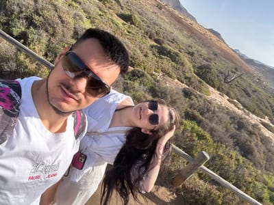 A selfie taken by a man and woman outdoors shows them standing on a hillside overlooking a dry, scrub-covered slope under a clear blue sky. The man, in the foreground, wears a white t-shirt with the text "LAUREN PAUSE JEANS" and sunglasses. The woman, to his right, wears a white top and sunglasses, with her hand resting on her head of long, dark hair. They appear to be on a hiking trail, with a wooden railing visible to their right. The terrain suggests a coastal or arid environment. This landscape is characteristic of the Cabo de Gata-Níjar Natural Park, located near the city of Níjar, Spain.