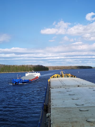 The image shows two barges on a body of water with a forested shoreline in the distance under a sky with scattered clouds. One barge, closer to the viewer, has a gray deck with evenly spaced manholes, and a yellow superstructure. A portion of blue railing is visible along its side. The second barge, further away, is blue and red with a gray cargo cover, and a white superstructure bearing the Cyrillic text "ОКСКИЙ 45 И." People can be seen on the side of the closer barge. The location is likely near Edoma, Russia, given the Cyrillic inscription and general landscape.