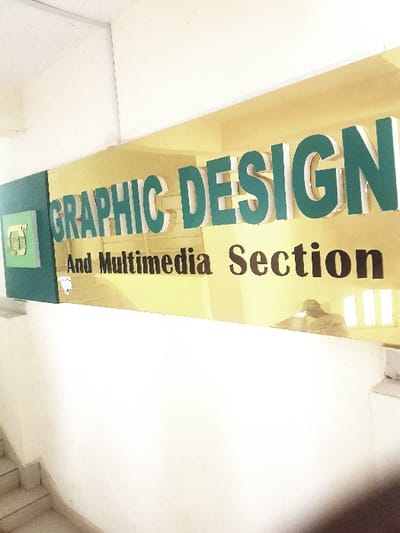 The image captures an indoor scene, primarily featuring a large rectangular sign mounted overhead, likely on a ceiling or high wall, viewed from a low angle. The sign is oriented vertically from the perspective of the viewer, meaning the text runs upwards.
The main part of the sign has a bright, light yellow or cream-colored background. On this background, large, three-dimensional letters spell out "GRAPHIC DESIGN." These letters are a prominent teal or dark green color with distinct white or light-colored edges, giving them a raised, block-like appearance. Below "GRAPHIC DESIGN," in smaller, darker, possibly brown or black text, it reads "And Multimedia Section."
At the very bottom of the sign (which would be the leftmost part if the sign were oriented horizontally), there is a smaller, dark green rectangular section, possibly featuring a subtle logo or symbol, though it's indistinct.
The surrounding environment consists of plain white or off-white walls and ceilings, creating a clean, bright, and somewhat stark atmosphere. In the upper right background of the image (or upper left if the text is read as intended from left to right), a bright rectangular light source, possibly a window or a large light fixture, is visible, casting a strong illumination that suggests a well-lit space. There's also a faint, blurry shape in that area, which could be a distant object or a person. The image has a slightly overexposed quality, particularly in the white areas, indicating bright lighting conditions. The overall impression is that of a sign marking a specific section or department within a building, such as a school, university, or office.