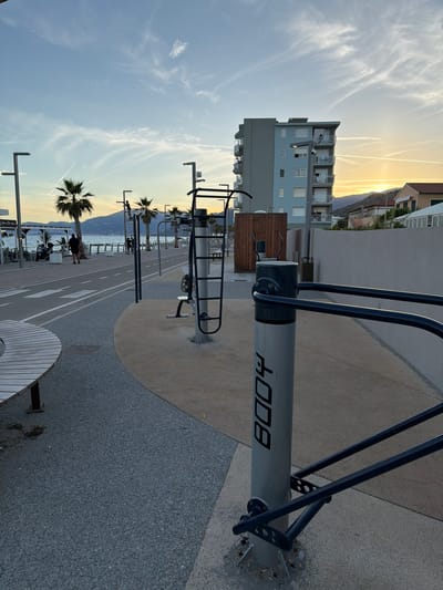 The image captures an outdoor exercise area with a promenade and the sea in the background, likely in Vallecrosia al Mare, Italy. The scene is illuminated by the soft light of either sunrise or sunset. In the foreground, a metal exercise station with parallel bars and a vertical support pole is prominently featured. The pole has the word "BODY" printed vertically in black capital letters. To the left, a curved wooden bench sits on a textured grey paved surface.
Further back, a paved path with white lane markings runs parallel to the sea, which is visible in the middle ground. A few palm trees are scattered along the promenade. In the background, a multi-story building with balconies and several smaller houses are visible against a backdrop of hills. The sky displays streaks of clouds and the sun is low on the horizon, casting a warm glow. Two individuals are walking on the promenade, and another appears to be interacting with an exercise apparatus. The overall environment suggests a public recreational space by the coast.