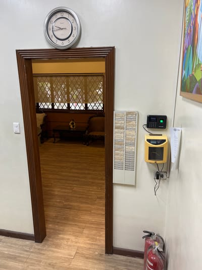 The image shows an interior scene with a doorway on the left leading into a room with wooden floors and furniture. On the wall to the right of the doorway, there are several items mounted. At the top, there is a round analog clock. Below the clock, there is a time clock machine. To the left of the time clock, there is a card rack filled with employee time cards. Further to the right, there is a piece of paper tacked to the wall. At the bottom right of the image, there are two red fire extinguishers. The overall environment appears to be an office or administrative area.