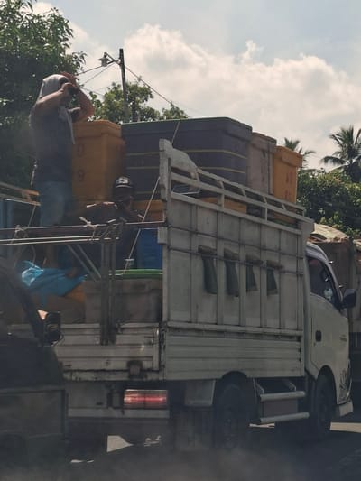 A blurry, medium shot captures a light-colored truck loaded with large yellow and purple containers driving on a road in Lhoknga, Indonesia. The truck has a white cab with a small dark shadow near the driver's side, suggesting a person is inside. In the bed of the truck, a person wearing a dark shirt and a head covering is visible. Another person wearing a dark hat is seated further back, possibly looking at their phone. Several large, rectangular containers are stacked on the truck's flatbed. The surrounding environment shows lush green trees and a cloudy sky with palm trees visible in the distance. The truck's brake lights are illuminated.