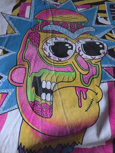 The image is a close-up shot of a t-shirt with a graphic print. The graphic depicts a stylized, somewhat grotesque, cartoonish face with exaggerated features, possibly inspired by the character Rick Sanchez from the animated series "Rick and Morty." The face is predominantly yellow, with green and pink details representing exposed brain matter and veins. The eyes are large and wide, with red veins visible. The mouth is open, revealing white teeth. Pink liquid is dripping from the mouth. The background of the graphic features blue star-like shapes and geometric patterns in pink and yellow. The t-shirt itself is white with colored stripes visible on the sides. The fabric of the t-shirt is textured and shows some creasing, indicating it is a wearable garment. There is no indication of any specific location in the city of Širvintos, Lithuania, or any other urban environment. The image focuses solely on the printed design on the clothing.