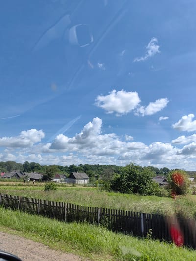 The image depicts a rural village scene under a bright blue sky with scattered white clouds. The foreground features a dirt path bordering a grassy area with a wooden fence running horizontally. Beyond the fence, a field is filled with tall grass and wildflowers, interspersed with several village houses with grey roofs. A dense line of green trees forms the backdrop, suggesting a forested area behind the village. In the distance, more houses are visible, some with chimneys. The sky is a dominant element, with noticeable contrails from aircraft. The scene appears peaceful and idyllic, characteristic of a rural landscape. The location is identified as Ilyanskiy selskiy Sovet, Belarus.