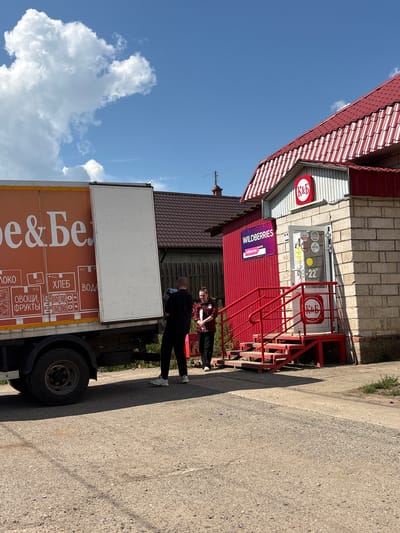 A delivery truck is parked next to a Wildberries and K&B store in Mysy, Russia. The truck has an orange side with white lettering and graphics indicating food items. Two people are standing near the truck and the store. One person, dressed in black, is carrying a bundle of items and appears to be interacting with the other person, who is wearing a red plaid shirt and black pants and is holding a phone. The K&B store has a red awning and red stairs leading to the entrance, with a sign displaying the K&B logo. The sky is blue with some clouds. The ground is asphalt and dirt.