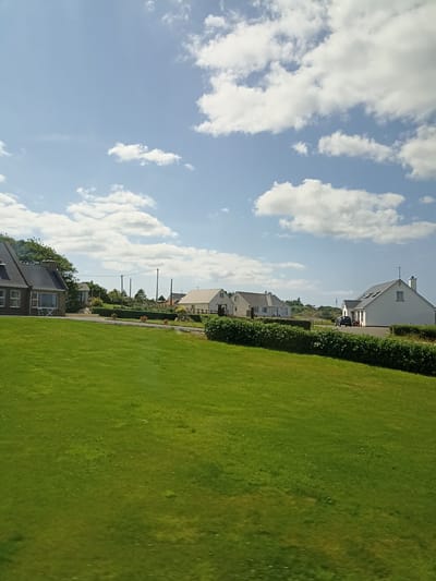 The image depicts a suburban residential scene in Belcruit Upper, Ireland. The foreground is dominated by a lush, green lawn that slopes gently upwards towards a row of houses and hedgerows. The houses are modern, with white or light-colored exteriors and pitched roofs. Some of the houses have chimneys, and several have visible windows.

Behind the houses, there are trees and foliage, and further in the distance, there are what appear to be telecommunication poles and possibly some fencing. The sky is a bright blue with scattered white clouds, suggesting a pleasant day. A dark-colored car is parked in the driveway of one of the houses. There are no people visible in the image. The overall impression is one of a quiet, well-maintained residential area.