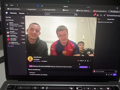 A MacBook Pro laptop displays a live Twitch stream originating from Krems an der Donau, Austria, as indicated by the channel name "manofkrems." The stream's video feed shows three young adult males in an indoor setting with light-colored walls. The individual on the left has short dark hair, a light mustache, and is smiling while wearing a dark hooded sweatshirt. The central individual wears glasses, a red and blue FC Barcelona football jersey, and is also smiling. The third individual, partially visible on the right, has dark hair, wears a black and white striped athletic shirt, and holds an object resembling a pipe or vape pen.
The Twitch interface indicates the stream is titled "Laf Moabet," categorized as "Just Chatting," and tagged with "Български" (Bulgarian). It shows a current duration of 0:02:18 and 0 viewers. The channel "manofkrems" has 0 followers. A "Stream Chat" panel on the right displays "Welcome to the chat room!" The left sidebar lists various "Live Channels" with viewer counts. The browser's tab bar includes open tabs for "Work," "Agent Orchestration," "Google Gemini," "Color Palette: #F7F6E...", "twitch.tv," "Claude," "New chat - GitHub Co...", and "Online Sportwetten in...". The bottom portion of the laptop, including the keyboard and trackpad, is visible.