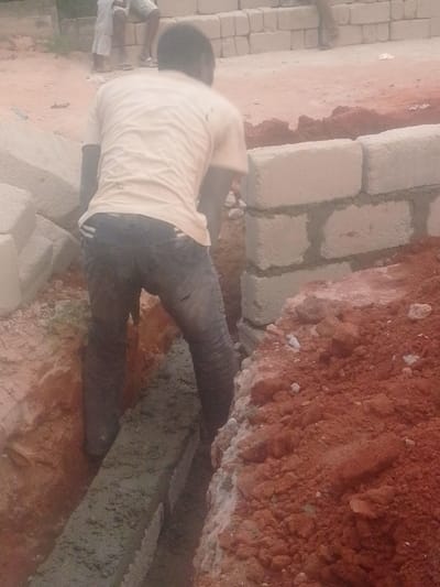 The image captures an outdoor construction scene, likely during the daytime. The foreground is dominated by a person, seen from behind, who appears to be engaged in manual labor. This individual wears a light-colored, possibly beige or pale yellow, short-sleeved t-shirt and dark trousers, which appear dirty with earth or cement. The person is bending forward, standing inside a narrow, deep trench that runs roughly vertically through the lower half of the frame.
At the bottom of this trench, a substantial layer of wet, grey concrete or cement is visible, suggesting that a foundation or footing is being poured or has recently been laid. The sides of the trench are rough, exposed reddish-brown earth.
To the right of the trench, a partially constructed wall made of light-colored concrete blocks (cinder blocks) is visible. Several courses of these blocks have been laid, cemented together. More stacked concrete blocks are visible further back and to the left of the main worker, indicating a supply of materials for the construction project. Piles of excavated reddish-brown soil are mounded along the edges of the trench, especially prominent in the lower right corner.
In the upper left background, two other individuals are partially visible. One is wearing white shorts and a white top, appearing to be sitting or crouching near a stack of blocks. The other figure is less distinct. The overall setting is a dusty, unpaved area. The image suggests an active construction site where a new structure, possibly a wall or building foundation, is being erected.