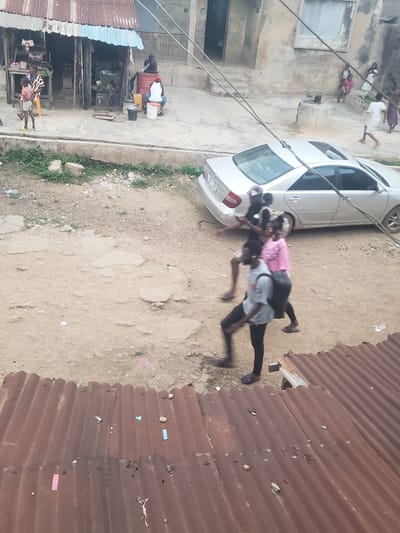 The image shows an outdoor scene in Alalubosa, Nigeria. A silver sedan is parked on a dirt surface. Several people are present, including a group of children on the left and two men engaged in what appears to be a physical activity, with one man carrying the other on his back. Buildings with corrugated metal roofing are visible on either side of the dirt area. There are additional people and objects in the background, suggesting a busy, densely populated environment.