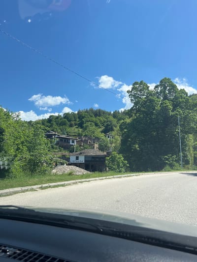 The image captures a view from inside a vehicle traveling along a road in Leshten, Bulgaria. On the roadside, a traditional Bulgarian building, possibly a guesthouse, is nestled on a hillside amidst dense greenery. A sign displaying the Cyrillic inscription "КРЪЧМА" is visible next to the building. The sky above is blue with scattered cumulus clouds. An overhead electrical line stretches diagonally across the sky. The road appears to be paved and well-maintained, running parallel to the vegetated hillside.