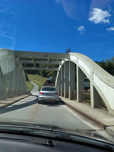 The image shows a silver Audi Q3 driving on a road under a concrete bridge. The car has a license plate that reads "PAN DA 34". The bridge has a series of arched concrete supports. Beyond the bridge, a winding road leads up a grassy hill with trees. The sky is blue with a few white clouds. The scene appears to be in a rural area. This is a view from inside a vehicle, looking forward through the windshield, with the dashboard visible in the foreground.