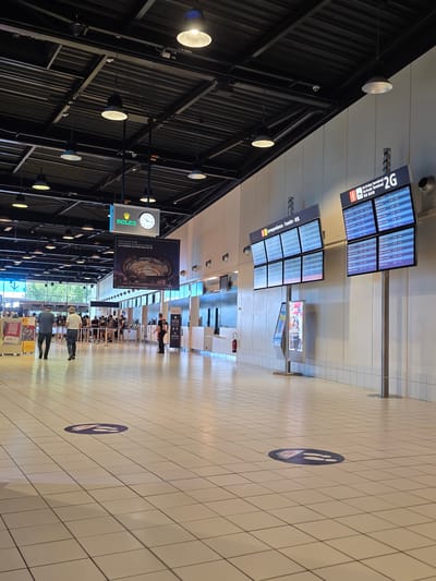 The image depicts the interior of a modern airport terminal, likely at Paris-Charles de Gaulle Airport (CDG) in Mitry-Mory, France, given the "Paris Aéroport" branding visible on a large advertisement. The scene is characterized by a bright, tiled floor and a high ceiling with industrial lighting and exposed infrastructure.
In the foreground, a circular social distancing marker with footprints is visible on the floor. Further back, several individuals are standing in line or walking through the terminal. Two men, one in a plaid shirt and the other in a white hat and jeans, are walking away from the camera towards the background.
Dominating the mid-ground are two large digital departure/arrival boards. The board on the left displays "Correspondances - Transfer" and shows flight information in multiple columns. The board on the right, labeled "Arrivées terminal 2G", also displays flight arrivals with destinations and times. A fire extinguisher is mounted on the wall to the left of the departure boards.
Further in the background, a large advertisement for a Rolex exhibition is suspended from the ceiling, featuring an image of the Panthéon. Above this, a prominent Rolex clock is also visible. Beyond the central area, more passengers are queuing, and counter-like structures suggest check-in or information desks. The overall environment suggests a functional, busy public space typical of an airport.