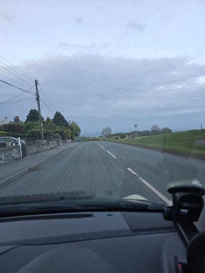 The image shows a view from inside a vehicle driving on a road in Halkyn, United Kingdom. The road has a single white line marking the lanes. To the left of the road, there are houses with stone walls and trees. A utility pole with wires is visible. On the right side of the road, there is a grassy verge. A speed limit sign is visible further down the road. The sky is overcast. A car mount is attached to the front windshield. The overall light level is low.