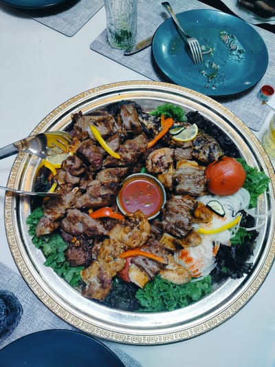 This image captures a close-up view of a large, ornate silver platter filled with a variety of grilled meats, vegetables, and garnishes. The platter is likely serving as a centerpiece for a meal, perhaps a celebratory feast or a family gathering. The grilled meats appear to be various cuts of lamb and chicken, some skewered and others arranged in generous portions. Colorful bell peppers, lemon slices, a whole grilled tomato, and fresh lettuce form the accompanying elements, adding visual appeal and freshness to the dish. A small bowl of red dipping sauce sits centrally within the platter.
In the background, partially visible elements suggest a dining setting. A blue plate with remnants of food and a fork indicates that diners have already begun eating. A tall glass of a beverage, possibly water with mint or a light-colored drink, rests on a gray placemat, as does another dark blue plate. The overall ambiance suggests a meal in progress, likely indoors given the lighting. The style of the platter and the food presentation could hint at a Central Asian culinary tradition, fitting with the provided location context of Aksukent, Kazakhstan. There is no visible text in the image. The image appears to be taken during daylight hours, but the exact time of day cannot be determined.