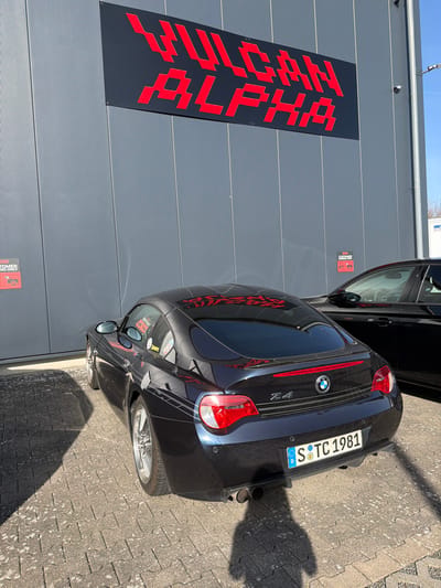 An outdoor scene in Meuspath, Germany, depicts a dark blue BMW Z4 Coupe (E86) parked on grey interlocking pavers in front of a grey industrial building. The vehicle is positioned facing away from the camera, revealing its rear and right side. It features silver multi-spoke wheels, a red decal on the rear windshield, multiple circular stickers on the rear quarter window, and white horizontal striping along its lower body. The German license plate reads 'S:TC 1981'. Dual exhaust pipes are visible on the rear bumper. To the right of the Z4, a black sedan, likely another BMW, is partially visible. The building's facade is composed of vertical grey panels and features a large black sign with 'VULCAN ALPHA' in red, pixelated lettering. A 'CUSTOMER PARKING ONLY' sign is affixed to the left side of the building, and a security camera is mounted on the upper right facade. A distinct human shadow is cast on the ground in the foreground, indicating the photographer's position. The environment is brightly lit by sunlight, casting sharp shadows.