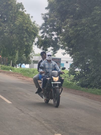 Two individuals are riding a black motorcycle on an asphalt road in Motala, India. The person in the front is wearing a checkered blue and white shirt and has a white cloth wrapped around their head, covering their face except for their eyes, which are visible behind dark sunglasses. The person behind them is wearing a dark jacket and grey pants. The motorcycle has the word "POLICE" written in white letters on the front fairing. The environment is characterized by trees and greenery on the sides of the road, with a building visible in the background. The sky is overcast.
