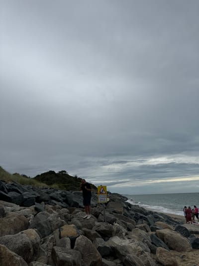 The image shows a rocky shoreline at Courtown, Ireland, under an overcast sky. A man in a dark t-shirt and shorts stands on the rocks, looking down. To his right, a yellow triangular warning sign with black pictograms and text in English and Irish, reading "Fan ar shiúl ó na cloché Keep off the rocks," is visible. Further down the beach, near the water's edge, a group of four women in casual clothing are standing. The sea is choppy with small waves breaking on the shore. The foreground is dominated by large, dark gray rocks. To the left, a grassy dune slopes upwards.