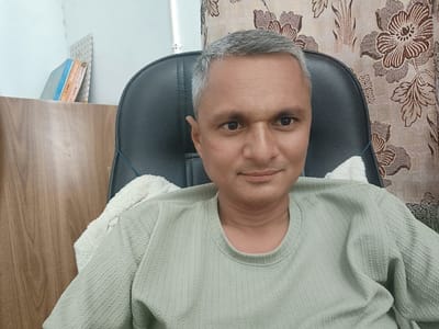In Dhandhuka, India, a man with short, graying hair and a mustache sits in a black office chair. He is wearing a light green, textured long-sleeved shirt. A white towel is draped over the back of the chair behind him. To the left of the man, a portion of a wooden cabinet is visible, holding two books. The background consists of a patterned curtain with floral designs in shades of brown and beige. The lighting appears to be natural, coming from the left.