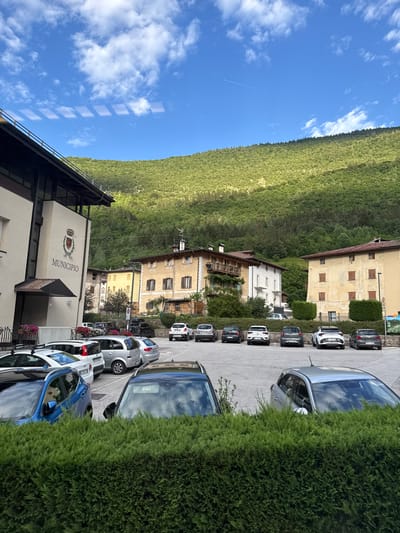 This media file shows a street scene in Spormaggiore, Italy. The image is oriented vertically, with the sky and a green, forested hillside occupying the upper portion, and a paved area with parked cars and buildings in the lower portion. Several cars are parked in a line along a paved surface. The buildings appear to be a mix of residential and commercial, with one building clearly marked "MUNICIPIO" with a crest above it, indicating it is the town hall. Some of the buildings have balconies and wooden shutters. The sky is a clear blue with scattered white clouds. The sun appears to be shining, suggesting it is daytime. The presence of a jet contrail in the sky indicates the time is likely midday or afternoon. The overall scene depicts a quiet, possibly rural European town on a clear day. There is no clear activity or event taking place, beyond the cars being parked.