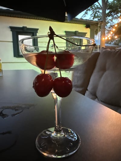 A close-up shot captures a clear glass cocktail with two cherries hanging from its rim, poised above a dark, reflective table. The cocktail glass is ornate, featuring a wide bowl atop a slender stem and base, filled with a light-colored liquid. The cherries, a deep red, are plump and glistening, their stems artfully arranged to suspend them. In the background, out of focus, is a building with a yellow facade and dark window trim, suggesting an outdoor setting, possibly a restaurant or cafe. String lights are visible further in the distance, adding a touch of ambient illumination. The overall scene evokes a sense of relaxed, evening ambiance. There is no indication of the city of Edineț, Moldova.