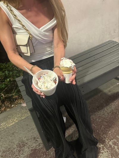 The image shows a person sitting on a gray metal bench, likely outdoors, holding both a cup of ice cream with a spoon and an ice cream cone. The ice cream appears to be vanilla and is topped with colorful sprinkles. The person is wearing a white top with a gold chain bag over the shoulder, and black pleated pants. The bench sits on what appears to be a concrete sidewalk with some greenery visible below the bench. The location appears to be outdoors, but there is no specific landmark or identifying information visible that would help determine the exact location in Boca Raton, Florida.