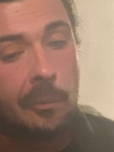 A blurry, low-resolution, close-up image depicts an adult male from the mid-chest up, located in Roanne, France. The subject has dark hair, a mustache, and a beard. His head is oriented slightly downwards and to the viewer's left, with his gaze directed downwards and his eyelids partially closed. His complexion shows reddish undertones. He is wearing a dark-colored garment, visible on the lower right portion of the frame. The background is a plain, light-colored, out-of-focus surface.