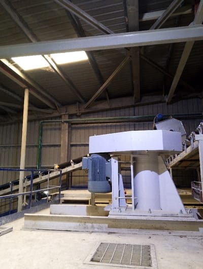 The image shows a large industrial machine, likely part of a cement factory, positioned in the foreground. The machine has a large, white, cylindrical body with a grey motor attached to one side. The machine is mounted on a concrete platform, and there is a metal grating next to it. The background is a large industrial space with a corrugated metal ceiling and walls. The building is brightly lit by fluorescent lights. The space is mostly empty, with only a few industrial components visible.  The location is an industrial facility in Pangasia, Bangladesh, with coordinates 22.693 latitude, 90.601 longitude. The image suggests an industrial environment, possibly a factory or a plant. There are no people present in the image.